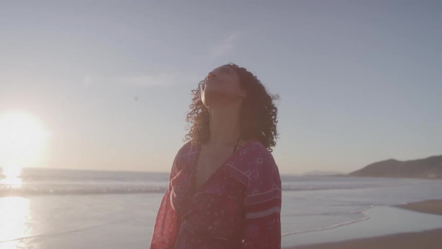 A young woman wearing a red dress stands on the beach at midday staring straight at the sky