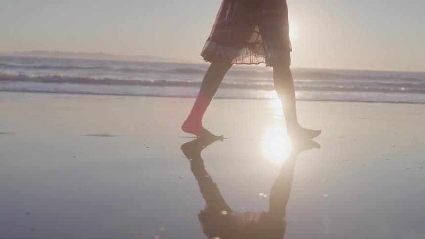 Legs of a woman walking on the wet sand of the beach with the sea and the sun in the background