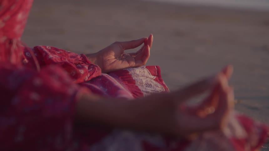 A person in a red patterned dress sits on a sandy beach meditating with their hands in a mudra position