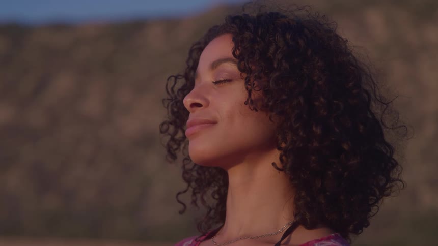 A young woman with curly hair stands with her eyes closed seemingly enjoying the peaceful outdoor environment