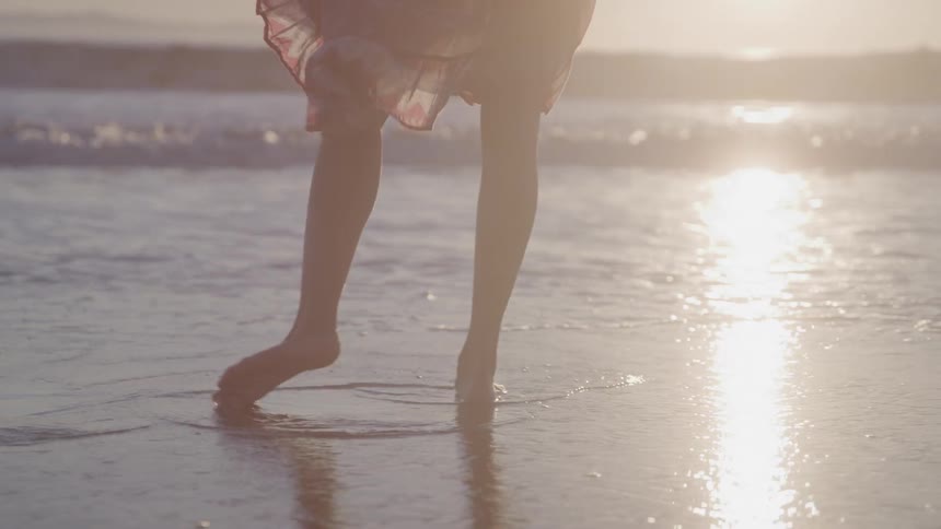 A persons bare legs and feet wade in the shallow water and wet sand on a beach at sunset