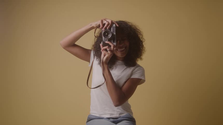 A young woman holding a digital camera sits in a room with yellow walls takes a snapshot and smiles as she looks at the photo