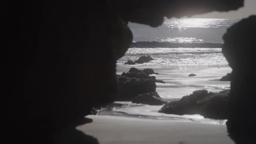 Through the dark silhouette of a rocky cave we can see the sun glints off the water as waves crash onto a sandy stonestrewn beach