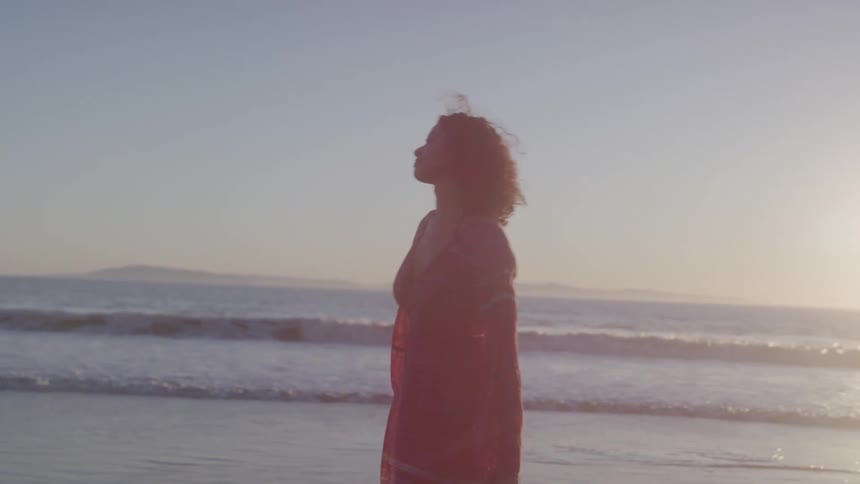 A woman with long curly dark hair wearing a flowing dress is standing on a sandy beach at sunset with her arms at her sides as she turns to look at the ocean