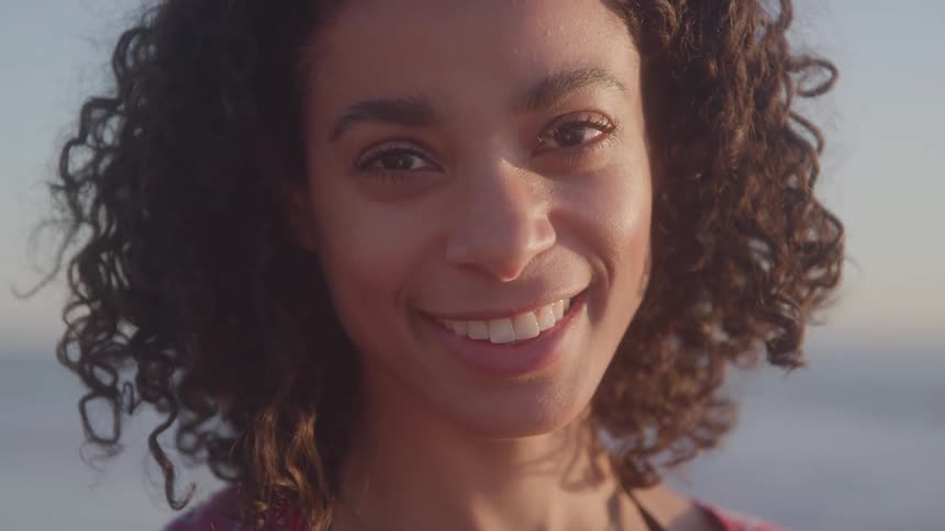 A woman with curly hair her face illuminated by the sun as she looks into the camera and smiles warmly