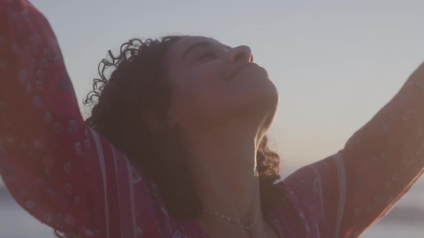 Curly haired woman with happy expression and open arms while standing on the beach