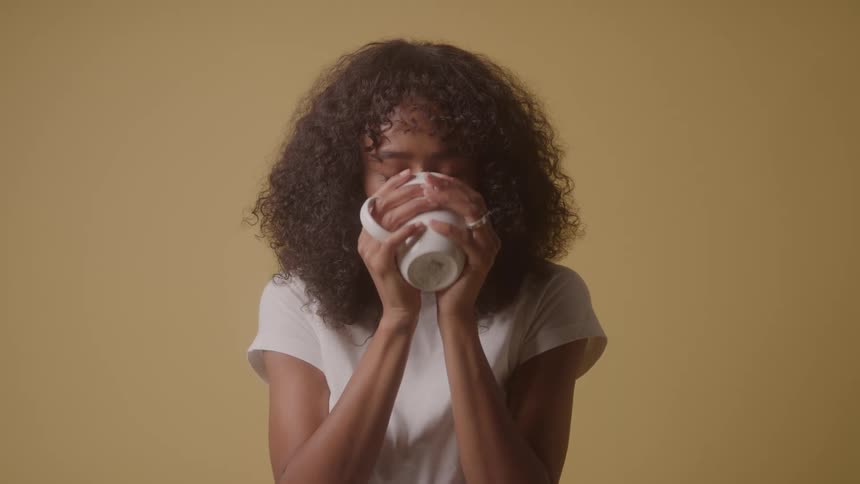 A woman with curly dark hair wearing a white shirt is standing in front of a tan wall holding a white mug with both hands as she smiles takes a drink from the mug and then smiles again