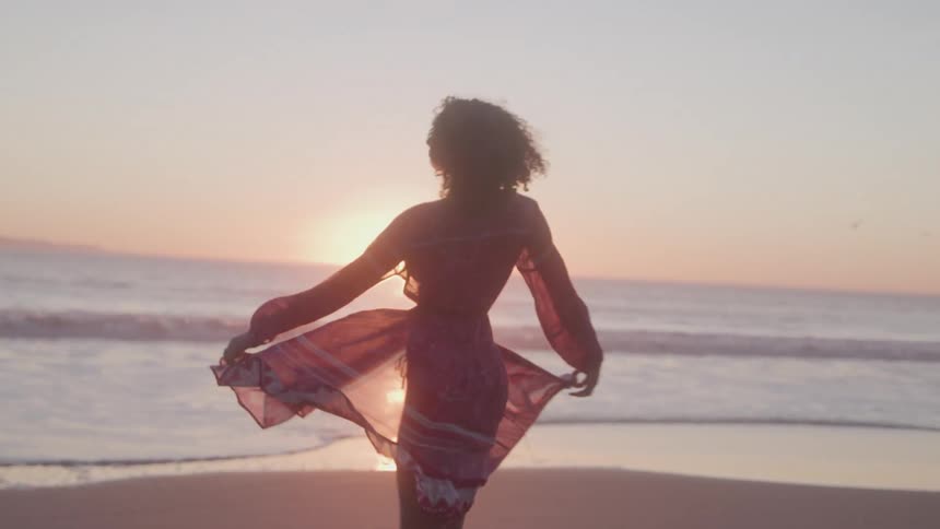 A woman in a light flowing dress stands on a beach at sunset spinning gracefully with her arms outspread as the waves crash gently behind her