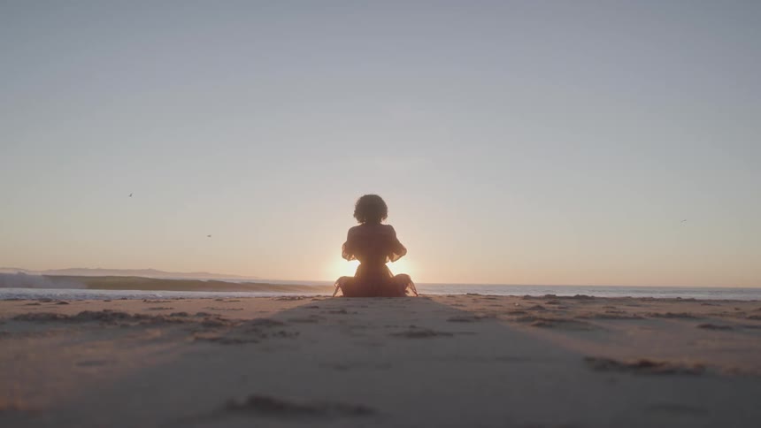 Young woman sitting on the beach sand meditating while watching the setting sun