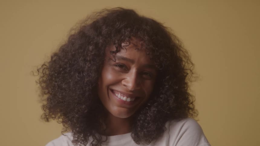 A young woman with big curly hair and a white tshirt smiles widely and laughs against a solid tan background
