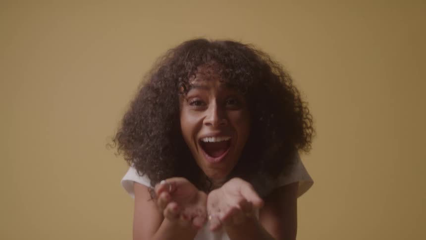 A young woman blows glitter from her palm as she laughs excitedly raising both hands in a room with yellow walls