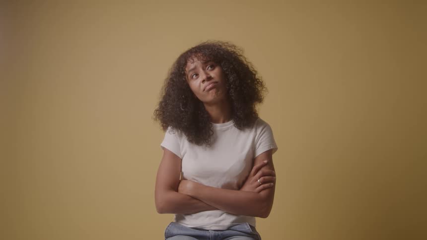 A frustrated young woman with curly hair sits with her arms crossed looking up and down with an irritated expression against a solid yellow background