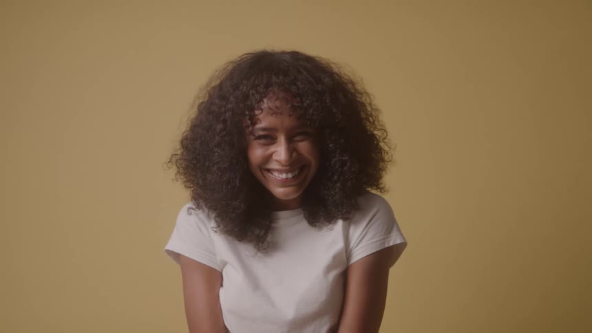 A woman with curly dark hair wearing a white shirt is standing in front of a tan wall looking directly at the camera with a big smile on her face before looking left and right with an even bigger smile