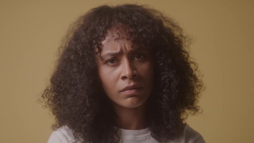 A young woman with long curly hair sits inside a room with yellow walls looking angry
