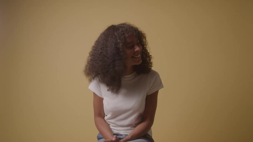 A woman with curly dark hair wearing a white shirt is sitting in front of a tan wall as she laughs and looks down while holding her hands to her mouth