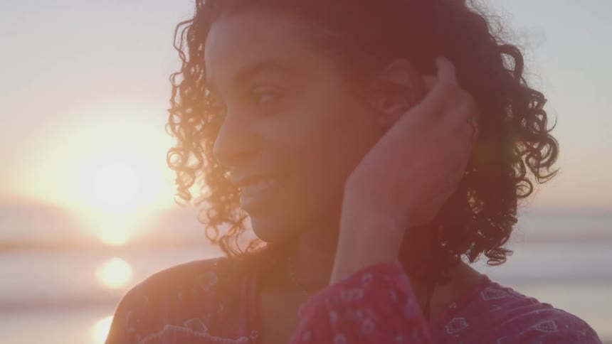 A woman with long curly dark hair is standing on a beach at sunset looking directly at the camera with a big smile on her face as she pushes some hair behind her ear