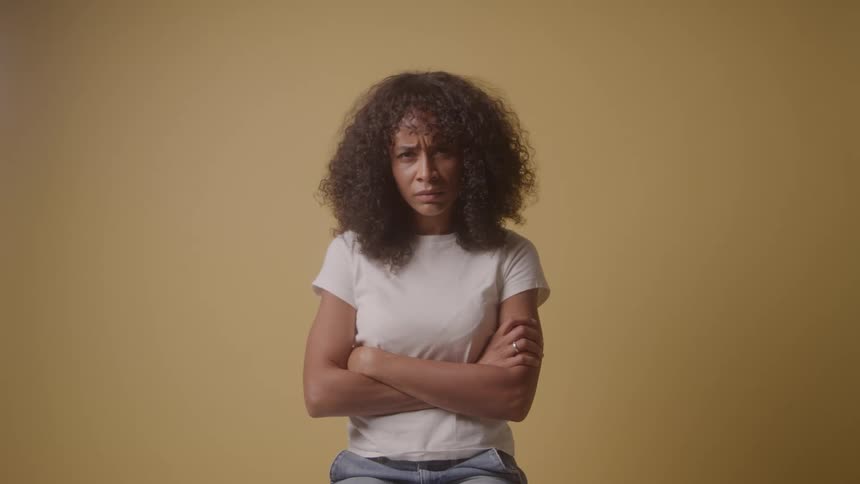 A young woman with curly hair sits inside a room with yellow walls folding her arms angrily