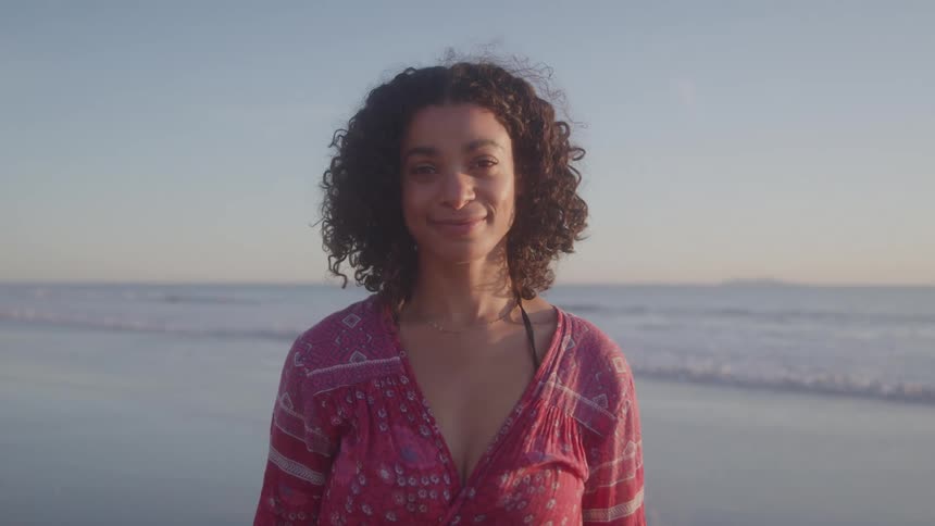 A smiling woman in a red patterned dress stands on a beach looking toward the ocean in the warm light of sunset