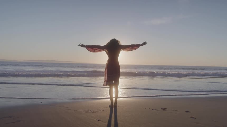 A woman with long wavy hair is standing on a sandy beach at sunset with her back to the camera and her arms stretched out to the sides as a sunbeam shines on the waves behind her