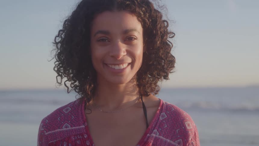 A young woman with curly hair stands on the beach wearing a red dress