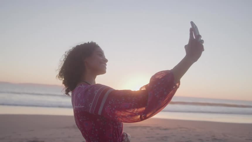 Young woman on the beach wearing a red dress taking a selfie with her phone at sunset