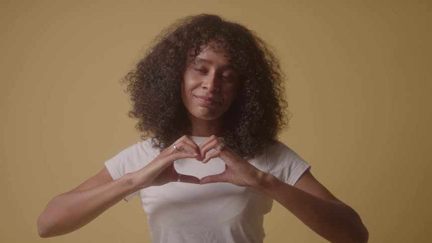 A young woman in a room with yellow walls making a heart sign with her hands while wearing a white tshirt