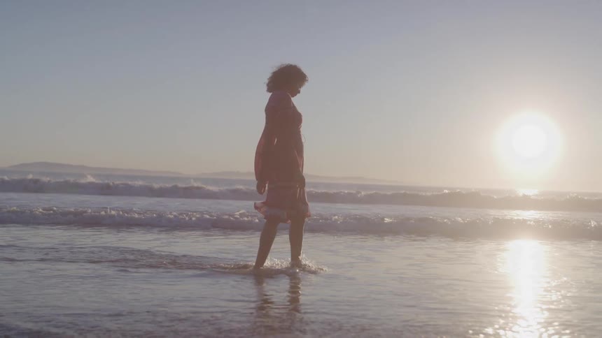 Woman in dress walking in beach water with morning sun on horizon