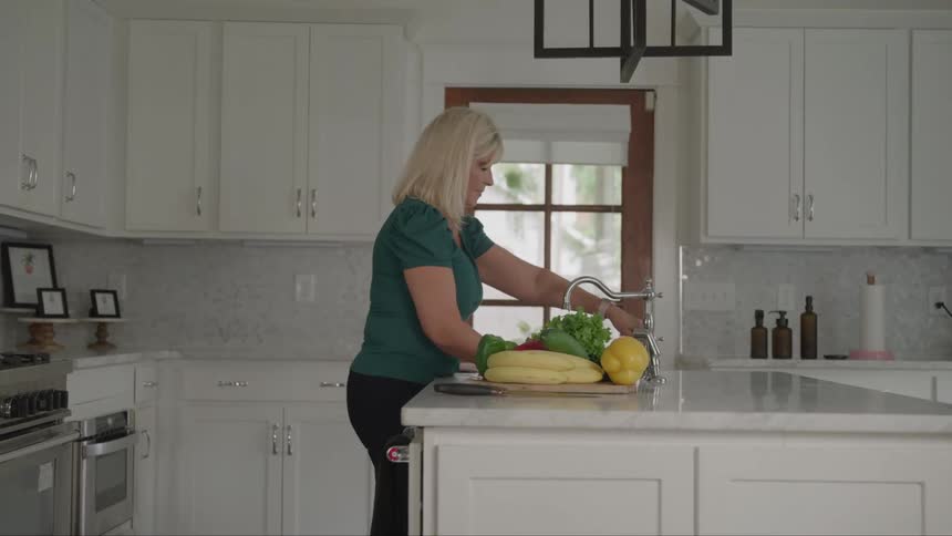A woman drinks water in a glass cup from a kitchen faucet indoors during daytime