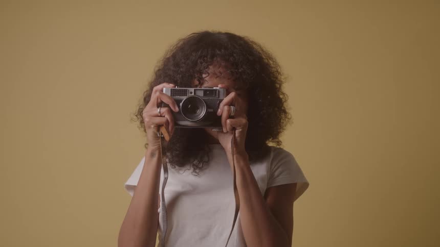 A young woman takes a photograph with a digital camera looks at the result and smiles inside a room with yellow walls
