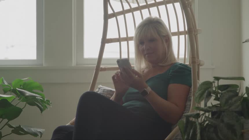 A woman sits indoors with potted plants on either side smiling as she scrolls through her phone at midday