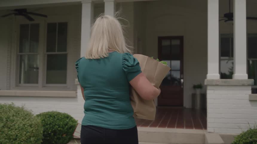 A woman carrying a grocery bag walks from her walkway to her front porch and then into her house at midday wearing a blue dress