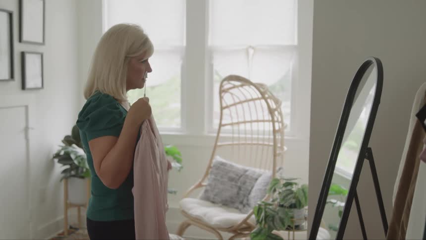 A woman picks up a dress from a hanger inside a room and checks its fit on herself in a mirror midday