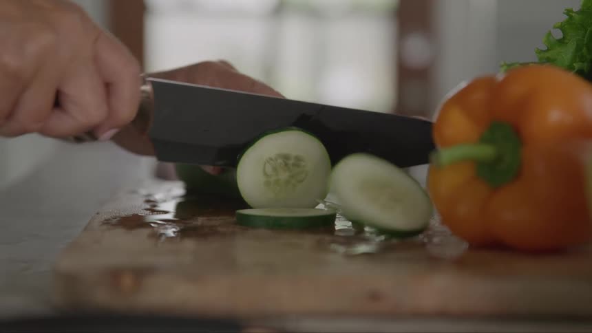 The video shows a pair of hands chopping a cucumber on a wooden cutting board with a large knife there is also a bell pepper and other vegetables visible on the board