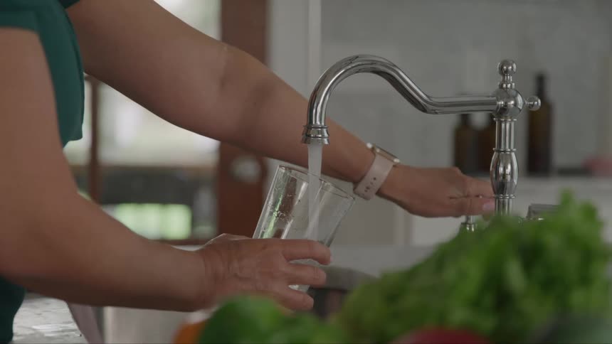 Person filling a glass with water from the tap in a kitchen with fresh vegetables on the counter