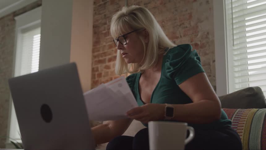 A blondehaired woman sits on a couch indoors sorting through documents with a laptop on the table in front of her and a coffee mug on the side at midday