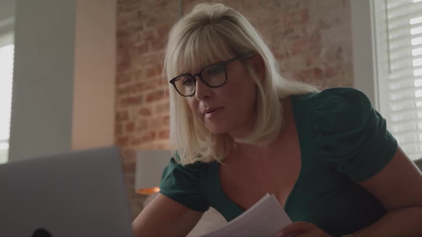 A blondehaired woman wearing glasses sits on a couch writing on a piece of paper and typing on a laptop at midday