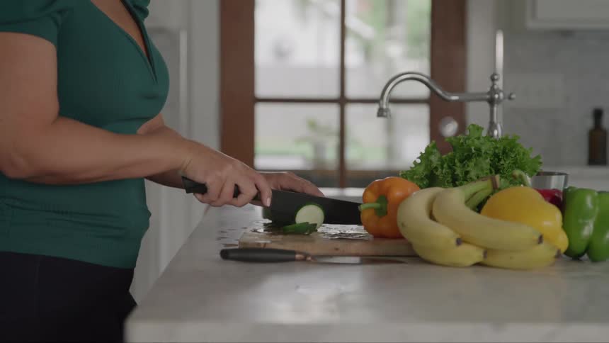 Person slicing cucumber in a kitchen with fruits and vegetables on the counter