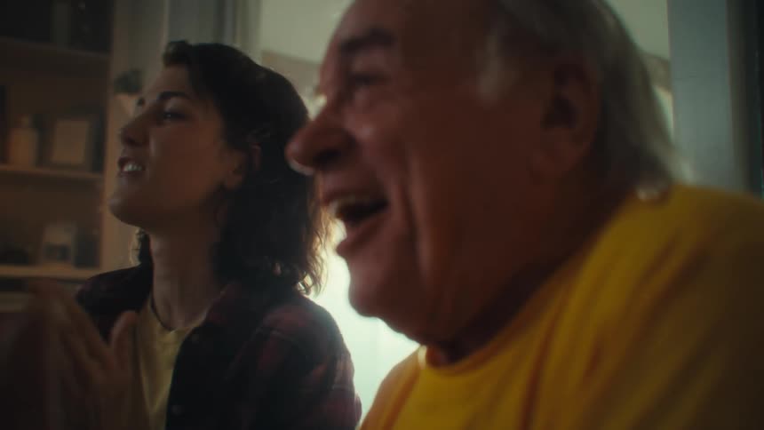 Close up of an elderly man and a young woman clapping and celebrating while watching tv in a living room during daytime