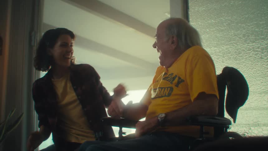An elderly man wearing a yellow tshirt that says army veteran sits in a wheelchair hugging a young woman with excitement as they highfive laughing and smiling at midday in the living room