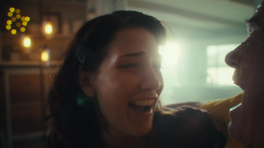A man with white hair wearing a yellow shirt and a woman with brown hair wearing a plaid shirt are sitting together as they both look at each other with happy expressions while the woman embraces the man