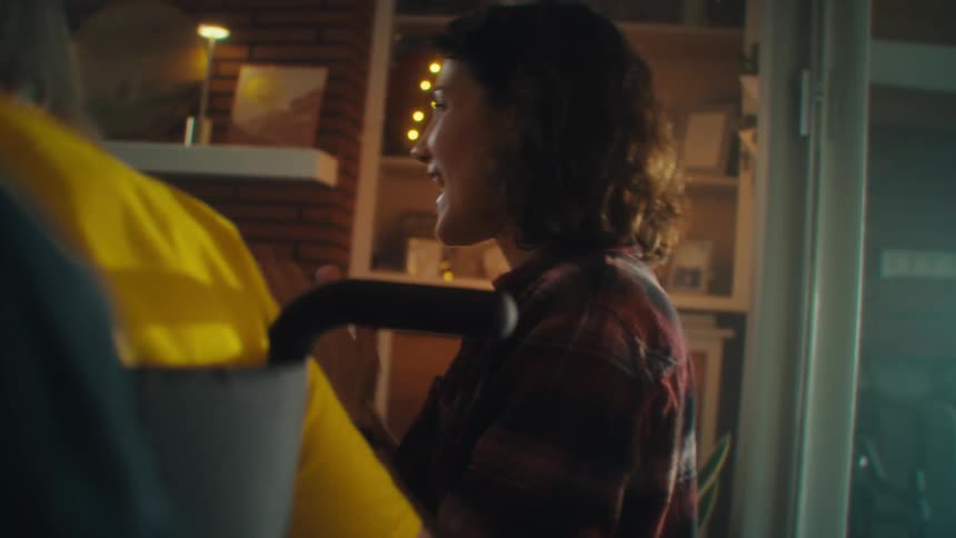 A woman with brown hair wearing a plaid shirt over a yellow shirt is celebrating and smiling next to a person who is sitting with their back to the camera