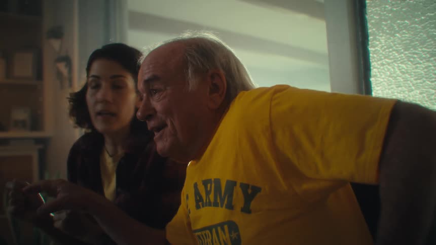 A man with white hair wearing a yellow shirt that reads us army veteran and a woman with brown hair wearing a plaid shirt are sitting together as they both look forward with excited expressions as they raise their arms in celebration