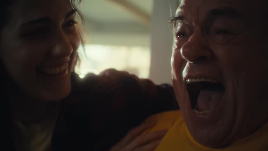 A video shows an elderly man and a younger woman sitting together both smiling and laughing the man wearing a yellow us army tshirt opens his mouth in a wide expression of joy and the woman laughs alongside him