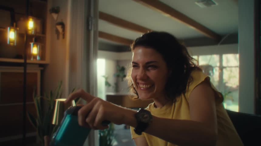 A woman with dark hair wearing a yellow shirt is sitting in a chair and playing a video game as she reacts with an excited and laughing expression