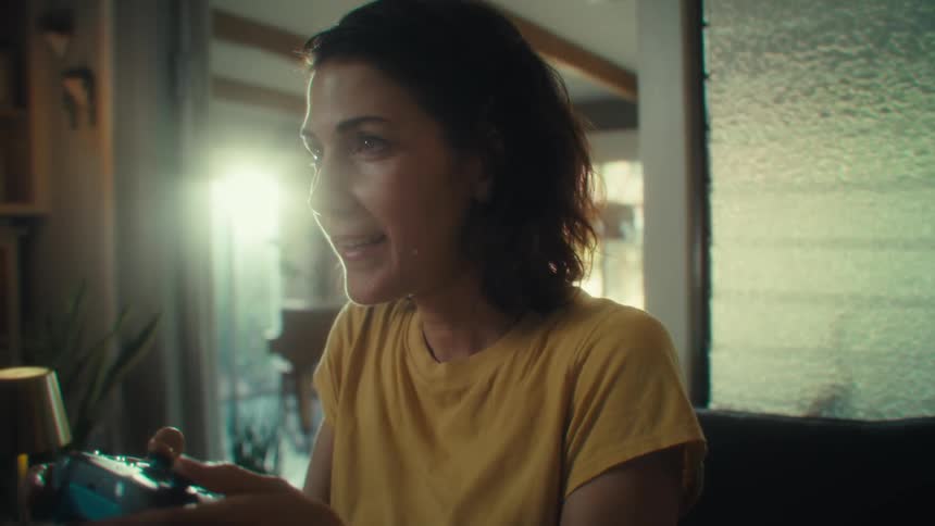 A woman with dark hair wearing a yellow shirt is sitting in a chair and playing a video game on a controller with an excited smile