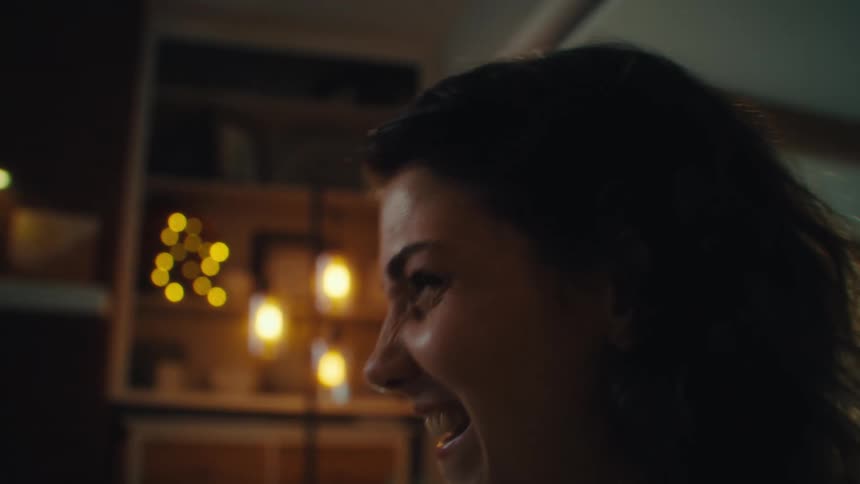A woman with dark hair wearing a yellow shirt is sitting in a chair and playing a video game as she laughs with an excited expression