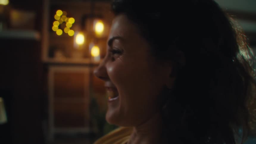 A close view shows a woman with dark hair and a light yellow shirt looking off to her right as her facial expression changes from serious to a big excited smile while playing video games
