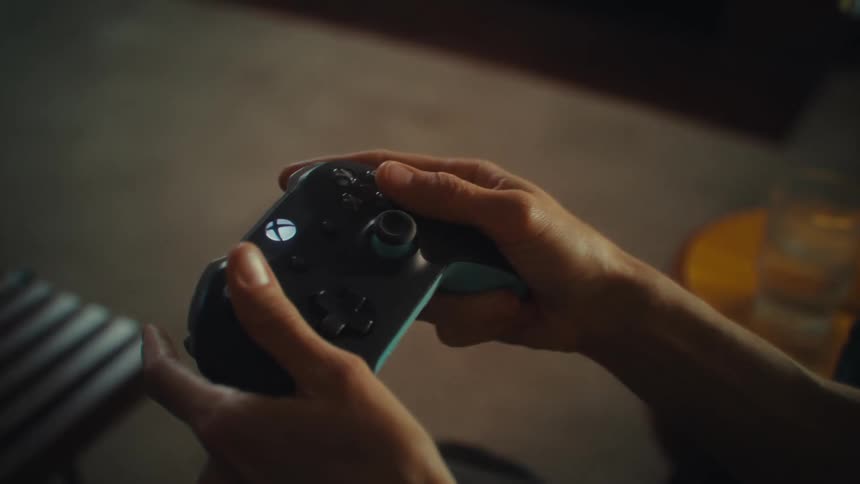 A close view of a womans hands shows her playing a video game on a black and gray controller with a glowing white symbol on it as she raises the controller in celebration