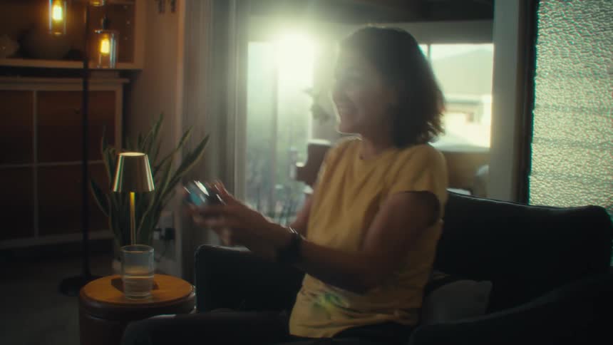 A young woman wearing a yellow tshirt sits on a chair in the living room gaming with excitement