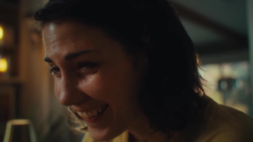 A woman with dark hair wearing a yellow shirt is sitting at a table and looking down at a smartphone as she begins to smile and laugh while holding a hand to her face